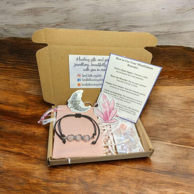 May include: A pink pouch with a black cord bracelet with clear crystal beads. The pouch is in a cardboard box with a card that says "How to Use Your Mindfulness Bracelet" and instructions on how to use the bracelet. The card also has a pink crystal graphic.