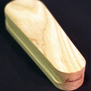 May include: A wooden, light brown, coffin-shaped box with a rounded top and a slightly curved bottom. The box has a red inscription on the bottom that reads "Yonecrafts".