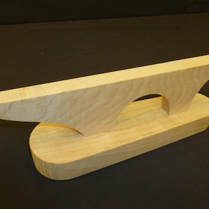 May include: A wooden clapper, a tool used in sewing and tailoring. The clapper is made of light-colored wood and has a rectangular base with a curved top. The top has a pointed end and a curved arch.