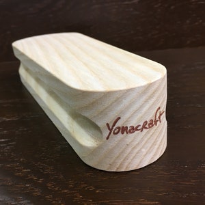 May include: A light wood climbing hold with a rounded shape and a small hole in the center. The hold is branded with the text "Yonacraft" in red.