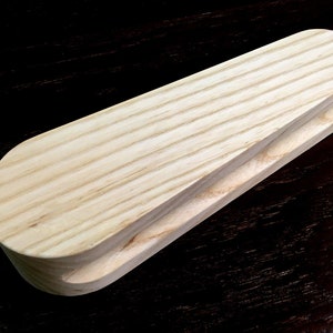 May include: A wooden tool for shaping fabric, with a rounded, slightly curved top and a groove on the bottom. The tool is made of light-colored wood with visible grain.