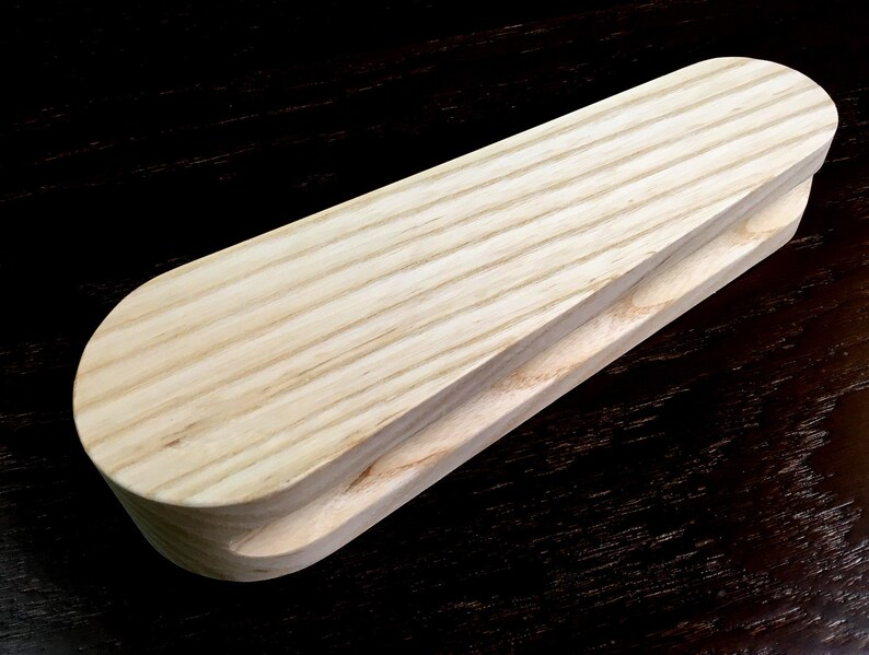 May include: A wooden tool for shaping fabric, with a rounded, slightly curved top and a groove on the bottom. The tool is made of light-colored wood with visible grain.