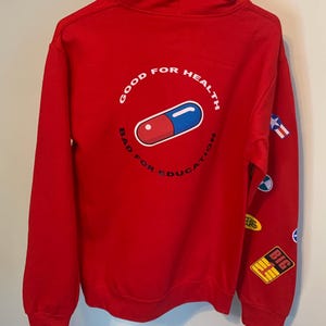 May include: Red hooded sweatshirt with a white circle graphic on the back that says "Good for Health Bad for Education" with a blue and red pill inside the circle. The sweatshirt has several small patches on the right sleeve.