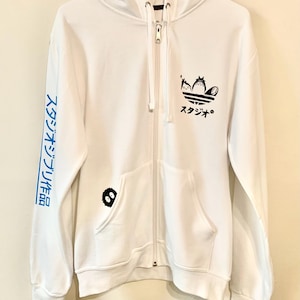 May include: White zippered hoodie with a black and white graphic of Totoro characters on the front. The graphic is a stylized Adidas logo with Totoro characters in place of the Adidas logo. The left sleeve has blue text in Japanese that reads "スタジオジブリ作品".