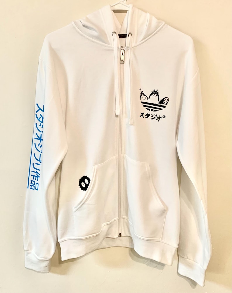 May include: White zippered hoodie with a black and white graphic of Totoro characters on the front. The graphic is a stylized Adidas logo with Totoro characters in place of the Adidas logo. The left sleeve has blue text in Japanese that reads "スタジオジブリ作品".