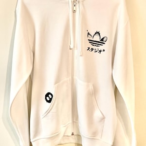 May include: White zippered hoodie with a black graphic of the Totoro characters from the Studio Ghibli film My Neighbor Totoro. The graphic is in the shape of the Adidas logo. The hoodie also has a small black Totoro graphic on the left pocket.