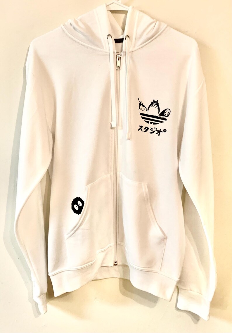 May include: White zippered hoodie with a black graphic of the Totoro characters from the Studio Ghibli film My Neighbor Totoro. The graphic is in the shape of the Adidas logo. The hoodie also has a small black Totoro graphic on the left pocket.