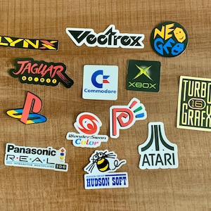 May include: A collection of colorful stickers featuring classic video game console logos, including Atari, Jaguar, and Xbox. The stickers are arranged on a wooden surface, showcasing a retro gaming theme.