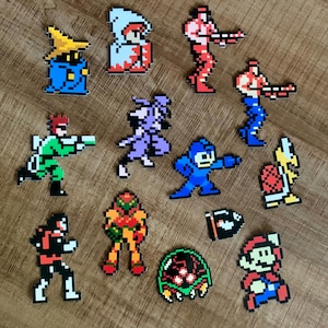 Könnte beinhalten: Eine Sammlung von Pixel-Art-Charakterfiguren aus klassischen Videospielen. Die Figuren umfassen Charaktere wie Mario, Mega Man und Samus, dargestellt in leuchtenden Farben. Die Figuren sind auf einer Holzoberfläche angeordnet.