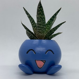 May include: A blue ceramic planter shaped like a smiling cartoon character with a large tongue sticking out. A small green succulent plant is growing in the planter.