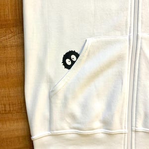 May include: White zippered hoodie with a black and white graphic of a cartoon character with two eyes peeking out of a pocket.