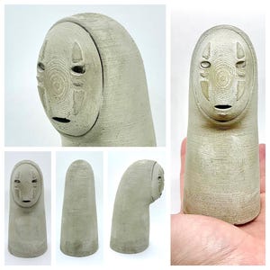 May include: A light gray, handcrafted sculpture of a character with a mask-like face. The sculpture has a cylindrical body and a rounded head with carved facial features. The surface has a textured, weathered appearance, suggesting a stone or concrete material.