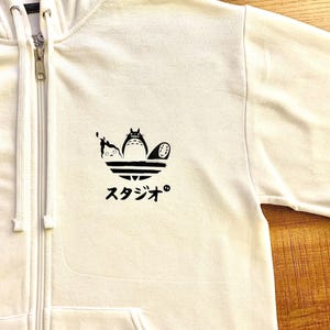 May include: White zippered hoodie with a black and white graphic of three characters from the animated film 'My Neighbor Totoro' on a stylized Adidas logo. The text 'スタジオ' is printed below the graphic.