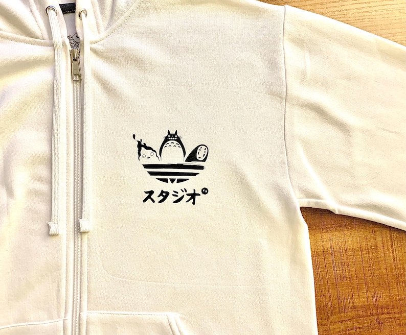 May include: White zippered hoodie with a black and white graphic of three characters from the animated film 'My Neighbor Totoro' on a stylized Adidas logo. The text 'スタジオ' is printed below the graphic.