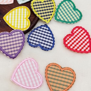 Könnte beinhalten: Eine Sammlung von herzförmigen Stoffaufnähern mit Vichy-Karo-Muster in verschiedenen Farben wie Rosa, Gelb, Blau, Grün, Schwarz, Lila, Orange und Rot, mit genähten Rändern, angeordnet auf und um eine kreisförmige Holzunterlage herum.