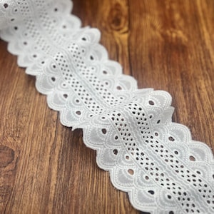 May include: White eyelet lace ribbon with a scalloped edge. The ribbon features a repeating pattern of small, circular cutouts and decorative stitching. The lace is displayed on a wooden surface.