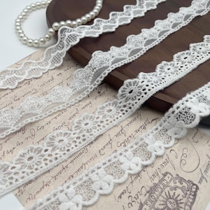 Puede incluir: Primer plano de varias cintas de encaje blanco con diferentes diseños florales y festoneados. Un collar de perlas y una bandeja de madera están en el fondo. Las cintas están sobre papel vintage con texto descolorido.