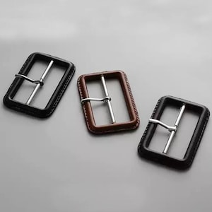 May include: Three rectangular belt buckles with rounded corners. Two buckles are black and one is brown. Each buckle features a silver-toned metal prong and a frame with visible stitching along the outer edge, providing a structured appearance.