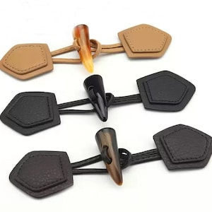 May include: Three toggle closures for garments, each featuring two pentagonal patches connected by a cord to a central horn-shaped toggle. The set includes one tan, one black, and one dark brown toggle closure, all with visible stitching.