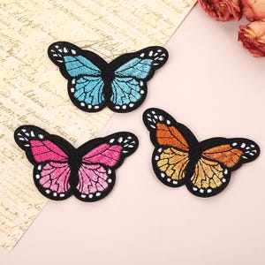 May include: Three embroidered butterfly patches featuring black borders and white-spotted wing edges. One patch has light blue wings, one has vibrant pink wings, and the third has orange and yellow wings with detailed black thread stitching.