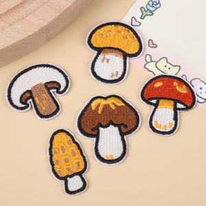 Puede incluir: Cinco parches bordados de setas con bordes negros. El conjunto incluye una seta blanca, una con sombrero amarillo, una con sombrero rojo, una con sombrero marrón y una seta alta y texturizada de color amarillo-marrón tipo colmenilla.