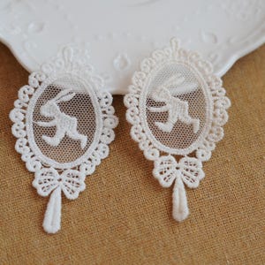 May include: Two white lace appliques with an oval design featuring a running rabbit. Each applique has a scalloped edge and a bow detail at the bottom. The appliques are on a tan fabric.