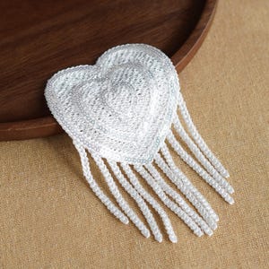 May include: A white heart-shaped patch featuring intricate embroidery and shimmering sequins. The heart is adorned with multiple long, dangling white beaded fringe strands that extend from the bottom edge, creating a textured and layered appearance.