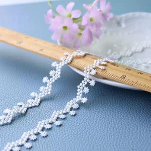 May include: A white floral lace trim ribbon featuring a repeating scalloped edge pattern. A wooden ruler is placed across the lace, showing measurements in inches, with the trim extending from the 6-inch mark to beyond the 9-inch mark.