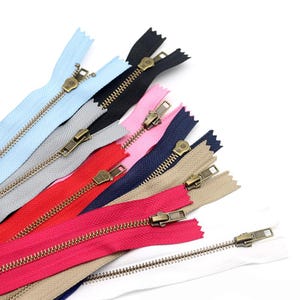 May include: A collection of ten fabric zippers in various colors including light blue, black, grey, pink, navy blue, red, beige, magenta, and white. Each zipper features a metallic bronze-toned pull tab and teeth, arranged in a fan-like pattern.