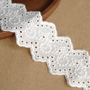 May include: Close-up of a white eyelet lace trim with a scalloped edge. The lace features a repeating pattern of stars and swirls, with small circular cutouts. The trim is placed on a brown surface, with a wooden bowl in the background.