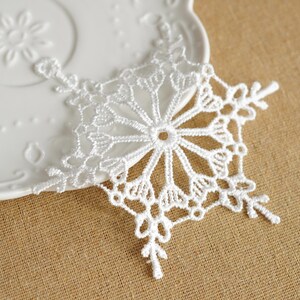 May include: A white crocheted snowflake ornament featuring intricate thread work with a six-pointed star design. The delicate lace-like pattern radiates from a central circular opening, showcasing detailed loops and branching points throughout the structure.