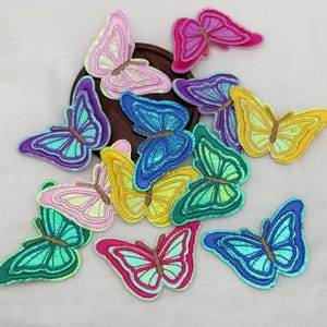 May include: A collection of embroidered butterfly patches in various iridescent colors including pink, purple, blue, yellow, and green. Each patch features detailed stitching and a shimmering finish, arranged in a scattered pattern on a light surface.