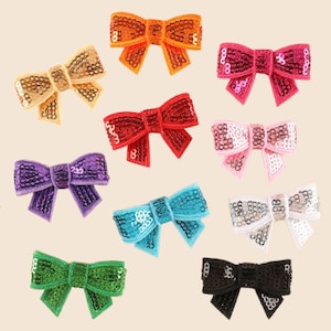 May include: A collection of ten decorative bows in various colours, including orange, gold, red, purple, green, pink, light pink, turquoise, white, and black. Each bow is covered in small, shiny sequins and features a central knot detail.