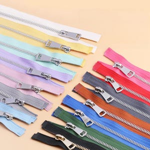 Puede incluir: Una colección de múltiples cremalleras en varios colores, incluyendo blanco, amarillo, azul claro, lavanda, rosa, gris, rojo, naranja, azul oscuro, verde y negro. Cada cremallera tiene dientes de metal plateado y un tirador metálico a juego.