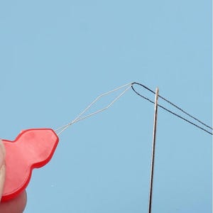 Puede incluir: Un enhebrador de agujas de plástico rojo con un fino bucle de alambre en forma de diamante que pasa por el ojo de una aguja de coser de metal plateado, con un hilo negro pasado a través del ojo de la aguja por el bucle de alambre.