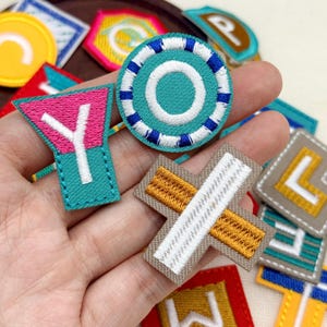 以下が含まれることがあります： 様々な文字や形が刺繍された布製ワッペンのコレクション。ターコイズブルーと白の円形の「O」、ピンクとターコイズブルーの「Y」、ベージュとゴールドの十字型のワッペンなど、様々な文字のワッペンが含まれています。