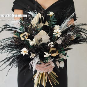 Pode incluir: Um buquê grande e dramático com flores e folhagens pretas, brancas e creme. O arranjo inclui capim-dos-pampas preto, margaridas brancas e anêmonas creme. O buquê é segurado por uma pessoa vestindo um vestido preto.