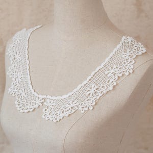 May include: A white, crocheted collar with a scalloped edge and a delicate floral pattern. The collar is displayed on a mannequin, showcasing its intricate lace design. This vintage-inspired accessory adds a touch of elegance.