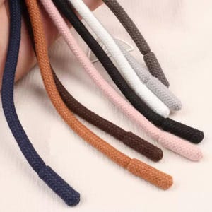 Puede incluir: Una colección de siete cordones trenzados en varios colores, incluyendo azul marino, tostado, rosa claro, negro, blanco, gris y marrón oscuro. Cada cordón presenta una punta trenzada a juego en el extremo para un acabado uniforme.