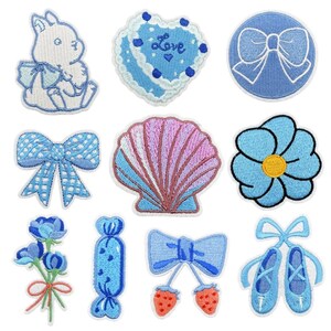 Può includere: Una collezione di undici toppe ricamate raffiguranti un coniglio bianco, un cuore blu con la scritta Love, un fiocco blu, un fiocco blu a quadri, una conchiglia rosa e blu, un fiore blu, un bouquet di fiori blu, una caramella blu, un fiocco con fragole e scarpette da danza blu.