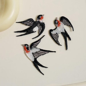 May include: Three embroidered swallow patches featuring black, white, and red thread. Each bird is depicted in a flying position with wings spread, showing detailed stitching on the feathers and a distinct red patch on the throat area.