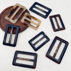May include: A collection of rectangular belt buckles in various sizes, featuring a smooth, marbled finish in shades of dark brown, black, and light beige. Each buckle has a central horizontal bar and two open rectangular slots for strap insertion.