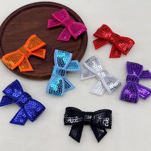Può includere: Una collezione di sette piccoli fiocchi in tessuto ricoperti di paillettes scintillanti, in una varietà di colori vivaci tra cui rosa acceso, arancione, rosso, azzurro, argento, viola e nero, disposti in modo sparso.