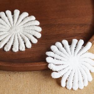 May include: Two white, embroidered flower appliques with a detailed petal design. The appliques are placed on a dark wooden tray, with a neutral fabric background.