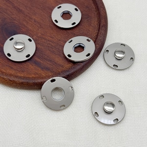 Peut inclure: Un ensemble de cinq boutons-pression métalliques circulaires de couleur argentée. Trois boutons présentent un dôme central surélevé, tandis que deux affichent un centre hexagonal plat et ouvert. Chaque pièce comporte quatre petites fentes.