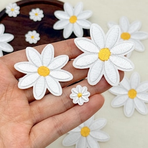 Puede incluir: Una colección de parches bordados de margaritas con pétalos blancos y un centro amarillo. Los parches varían en tamaño, con algunas flores más grandes y varias más pequeñas, todas mostrando costuras detalladas y un diseño circular limpio.