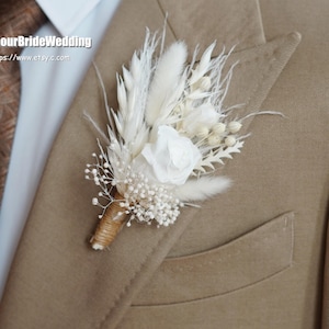 Op de afbeelding: Een witte gedroogde bloemen boutonnière met een witte roos, pampasgras en baby's breath. De boutonnière is vastgemaakt aan een bruin colbert.