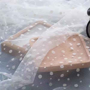 May include: A sheer white tulle fabric featuring a repeating pattern of small, opaque white polka dots. The lightweight, semi-transparent mesh material is draped over a light-coloured wooden tray, showcasing the delicate texture and dot design.