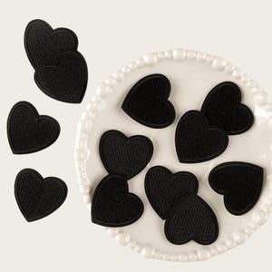 Puede incluir: Una colección de parches bordados en forma de corazón negro. Varios parches están dispuestos sobre un plato circular blanco con borde de cuentas, mientras que otros tres parches de corazón negro se colocan por separado a la izquierda del plato.