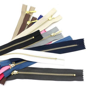 May include: A collection of fabric zippers arranged in a fan pattern, featuring various colors including black, white, navy blue, gray, brown, and pink. Each zipper has metallic gold-toned teeth and a matching gold-toned metal slider pull.